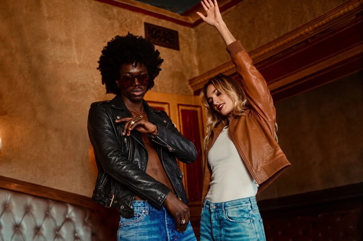 BAGGATTI PARIS campaign photograph: two models in a dimly lit Parisian interior, one in a black leather biker jacket and sunglasses, the other in a tan leather jacket over a white top with denim